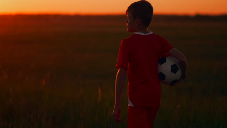 The-boy-is-on-the-field-with-the-ball-in-his-hands-looking-at-the-sunset-and-dreaming-of-a-football-career.-the-camera-follows-the-boy